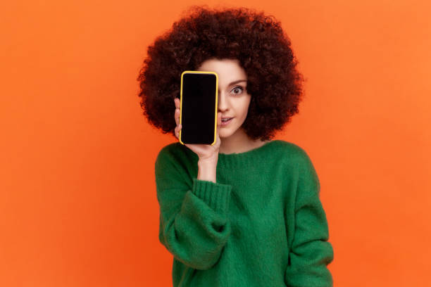 Attractive young adult woman with Afro hairstyle wearing green casual style sweater covering eye with cell phone with empty display. stock photo