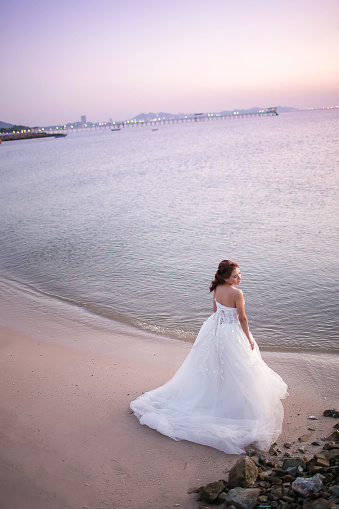 Attractive young bride wear wedding dress and white veil, stand alone in the Sea beach with rim light from the sun. bride in the Sea beach concept. image for background, copy space, objects fashion