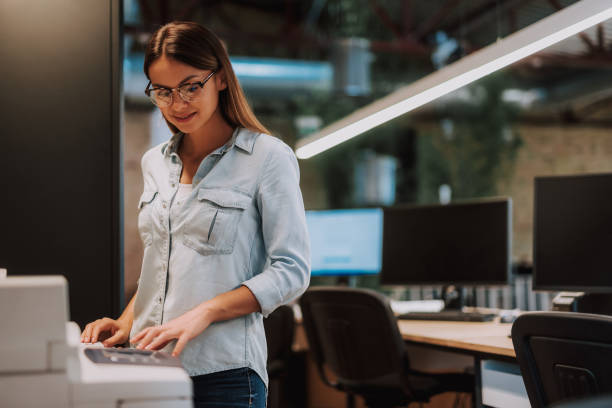 Attractive young lady in glasses working at modern office Waist up portrait of charming woman standing near photocopier and smiling. Desk with computers on blurred background xerox photocopy machine stock pictures, royalty-free photos & images