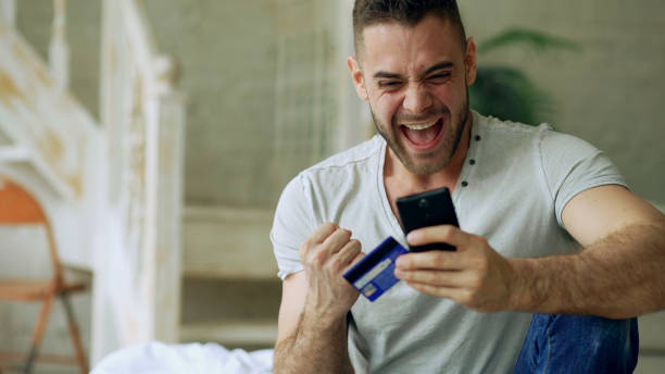 Attractive young man with smartphone and credit card shopping on the internet sit on bed at home stock photo