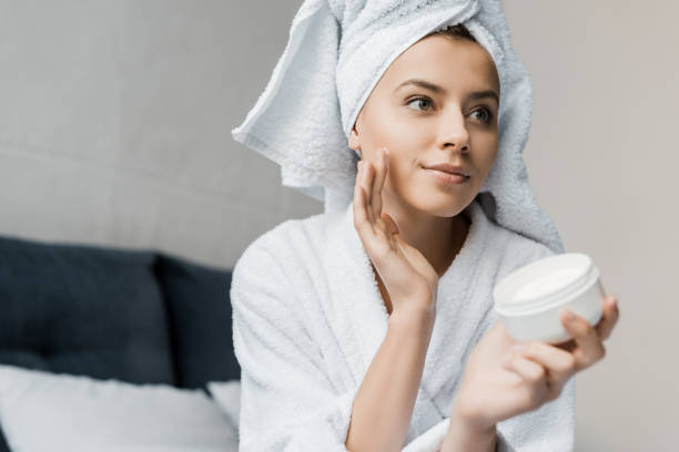 attractive young woman in bathrobe and towel on head applying moisturizing cream on face attractive young woman in bathrobe and towel on head applying moisturizing cream on face applying face cream stock pictures, royalty-free photos & images