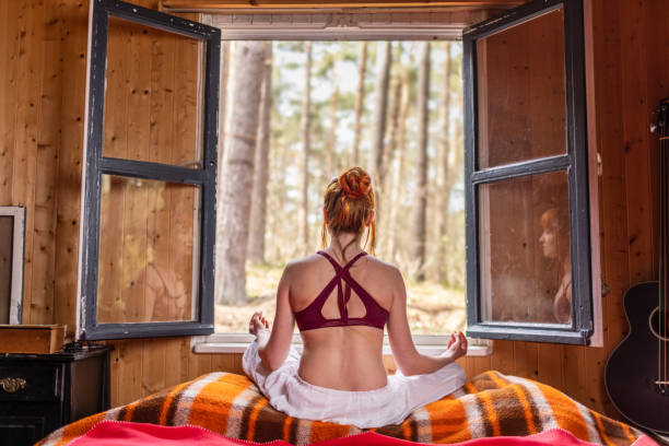 Attractive young woman meditating in bedroom with open window to nature and forest trees stock photo
