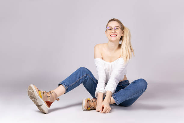 Attractive young woman posing over grey background. Studio shot of gorgeous young blonde woman with straight hair wearing off shoulder crop top sweater and high waisted denim shorts. Gray isolated background, copy space, close up. Ugly sneakers concept ugly skinny women stock pictures, royalty-free photos & images