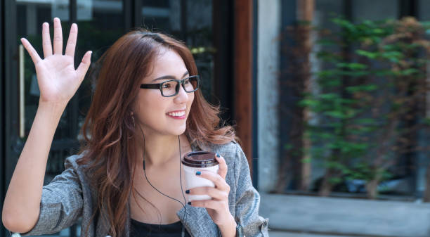 Attractive young woman sitting outdoor in cafÃ© while waving hand saying hello her friend. Business woman wear glasses holding cup of coffee while waving hands greeting her friend say hi or goodbye.  wave goodbye asian stock pictures, royalty-free photos & images