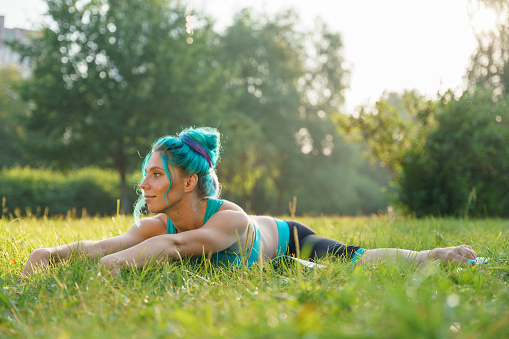 attractive young woman with a turquoise hair in a turquoise yoga outfit doing yoga on a grass in a park, during sunrise, in a beautiful sunlight, starches forward in a transverse twine and smiles, she is calm, enjoying her practice, not looking in a camera, we can see her profile