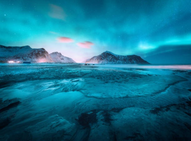 aurora borealis boven de besneeuwde berg en het zandstrand in de winter. noorderlicht in de lofoten eilanden, noorwegen. sterrenhemel met polaire lichten. nacht landschap met aurora, bevroren zeekust, stadslichten - northern light stockfoto's en -beelden