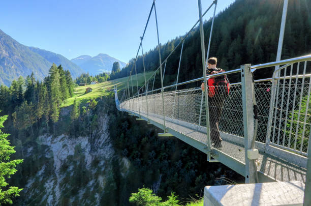 Austria's longest pedestrian suspension bridge in Holzgau Bridge of Holzgau HOLZGAU, AUSTRIA 18 June 2017: A man with a backpack walks over a suspension bridge in Holzgau, Austria. The village of Holzgau is famous for Austria’s longest (200m) pedestrian suspension bridge that opened in 2012, over Höhenbach Canyon, connecting Gföllberg and Schiggenberg. lechtal alps stock pictures, royalty-free photos & images