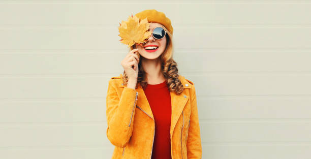 Autumn mood! happy smiling woman holding in her hands yellow maple leaves hiding her eye over gray wall background stock photo