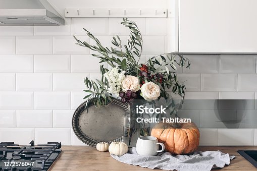 istock Autumn still life composition in rustic eclectic kitchen interior. Cup of coffee, vintage silver tray and floral bouquet. Wooden table background with pumkins. Thanksgiving, Halloween concept. 1272576047