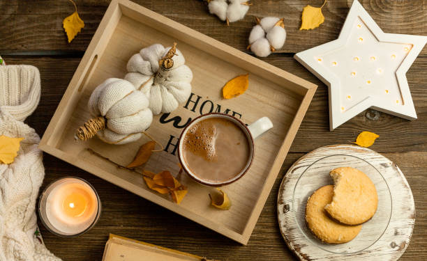 Autumn still life of a wooden tray, white knitted handmade pumpkins, yellow leaves, a mug of cocoa, coffee or hot chocolate, lit candles, round cookies. Warm home comfort concept stock photo