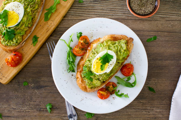 Avocado toast with eggs and roasted tomatoes Avocado toast with roasted tomatoes and eggs, shot from above on a white plate on a rustic wooden table. 
Served with fresh arugula and sprinkled with parsley and black pepper. 
Stock photo. avocado toast stock pictures, royalty-free photos & images