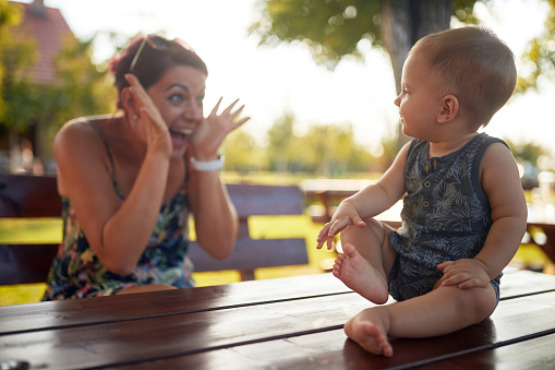 A baby enjoying sitting on a table and playing with mother in the yard on a beautiful weather. Farm, family, countryside, summer