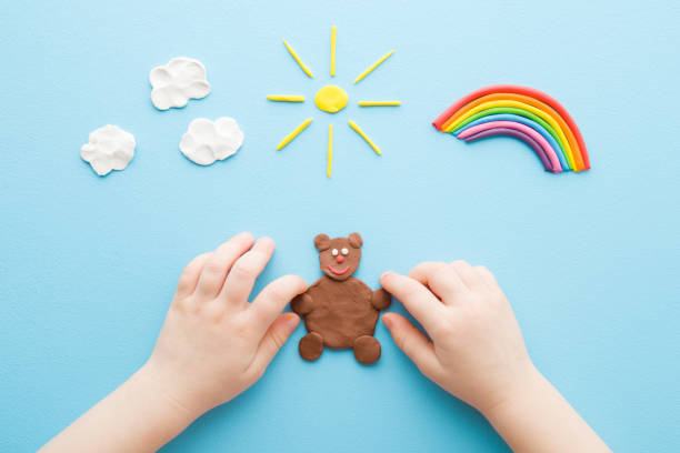 Baby hands making brown bear from modeling clay. Colorful rainbow, sun and white clouds on light blue table background. Pastel color. Closeup. Point of view shot. Toddler development. Top down view. stock photo