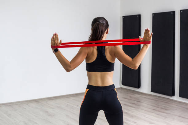 baksidan porträtt av sportig kvinna klädd i svart topp och leggings utför övningar för musklerna i rygg och händer, träning med motstånds bandet på vit bakgrund. - träningsgummiband bildbanksfoton och bilder