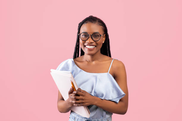 Back to school. Young black female student with book and notebooks smiling at camera on pink studio background stock photo
