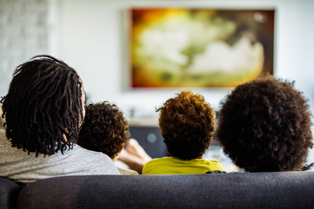 Back view of a family watching TV in the living room. Rear view of black family relaxing on sofa while watching a movie on TV at home. watching tv stock pictures, royalty-free photos & images
