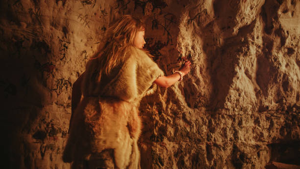 back view of a primitive prehistoric neanderthal child in animal skin draws animals and abstracts on the walls at night. creating first cave art with petroglyphs, rock paintings illuminated by fire. - fire caveman imagens e fotografias de stock