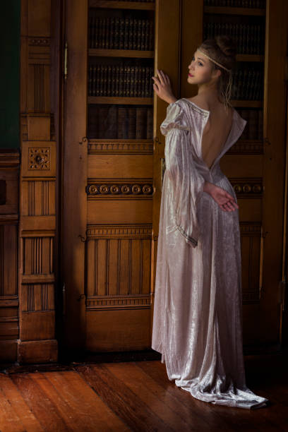 Back view of a young Caucasian woman in 20s wearing a long cream gown unzipped in the back leaning against a carved wooden bookshelf door in a study or library in a Gothic styled interior and looks over her shoulder with romantic longing Back view of a young Caucasian woman in 20s wearing a long cream gown unzipped in the back leaning against a carved wooden bookshelf door in a study or library in a Gothic styled interior and looks over her shoulder with romantic longing romance book cover stock pictures, royalty-free photos & images
