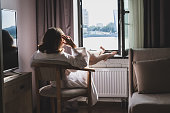 istock Back view of beautiful young woman in bathrobe sitting on armchair and enjoying near open window at sunny morning. 1362901302