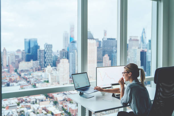 Back view of business woman sitting at panoramic skyscraper office desktop front PC computer with financial graphs and statistics on monitor. Analysis of digital market and investment in block chain stock photo