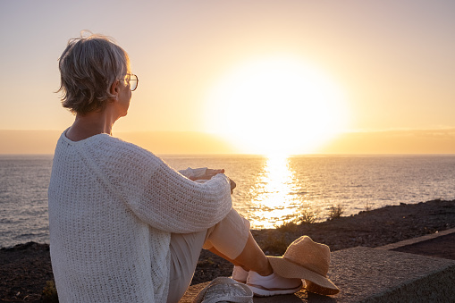 Back view of relaxed and romantic senior woman or pensioner sitting admiring the sea at sunset light looking at the horizon - old female  outdoors enjoying vacations