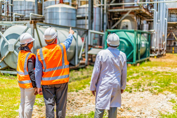 Back view of successful older business owner or manager in discussion with employees on site at a chemical plant. Diversity of ethnic background, Caucasian, African American and Indian. age and gender. All wearing appropriate PPE, Personal Protective Equipment. Non urban location. Outdoors. oil refinery factory stock pictures, royalty-free photos & images