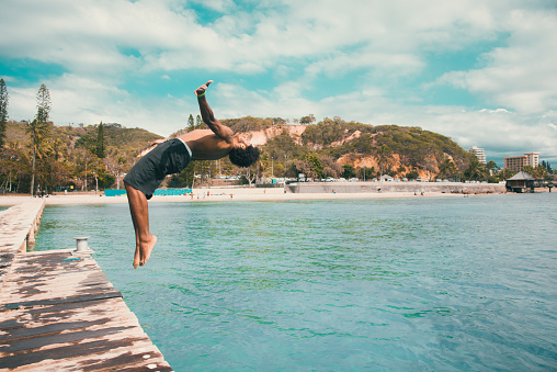Young pacific islander from the local kanak people tribe, the original inhabitants of New Caledonia jumping from wooden jetty with a backflip into the turquoise lagoon at the Beach of Noumea, New Caledonia. Real Native People Portrait. Noumea, New Caledonia, South West Pacific Ocean