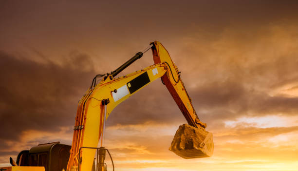 Backhoe working by digging soil at construction site. Closeup hydraulic arm and bucket of backhoe with scoop of soil against yellow sunset sky. Excavator digging on dirt. Trenching machine. stock photo
