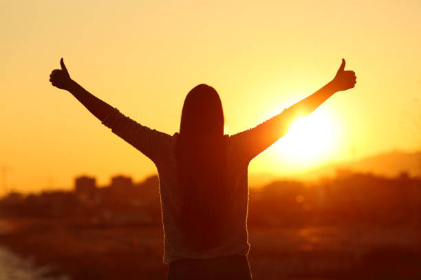 Backlight of a woman raising arms with thumbs up Back view backlight silhouette of a woman raising arms with thumbs up to the sun at sunset attitude stock pictures, royalty-free photos & images
