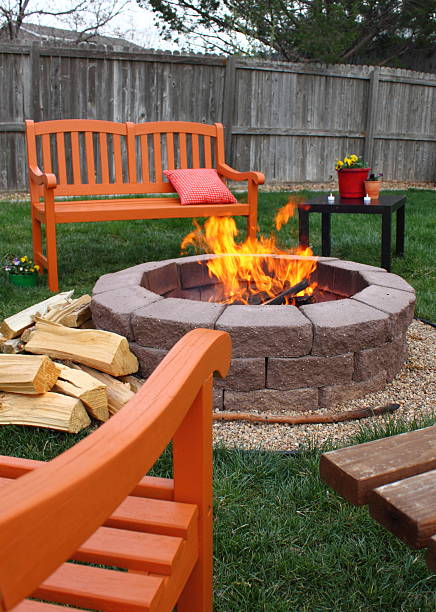 Backyard fire pit A charming back yard fire pit, surrounded by brightly colored orange chairs and benches. Around the flaming fire pit is a bright colored pillow, flower pots, and a pile of split wood, waiting to be burned. In the background is a weathered fence. Colors of green, brown, orange, red. fire pit stock pictures, royalty-free photos & images