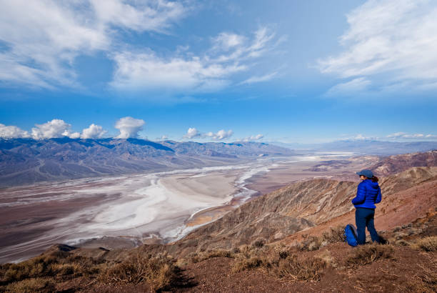 Hiker at Dante's View Death Valley National Park, California, USA - February 16, 2019: Badwater Basin is the lowest point in North America at 282 feet below sea level. Periodic rainstorms flood the valley floor but soon evaporate because of the intense heat, leaving behind a pattern of crystals on the salt pan. Average annual rainfall in Badwater Basin is 1.9 inches a year. According to legend, Badwater gets its name from a mule that refused to drink the salty water. This woman hiker is taking in the view of Badwater Basin from Dante's View at 5,476 feet above sea level. Dante's View is about 16 miles south of Furnace Creek. jeff goulden mojave desert stock pictures, royalty-free photos & images