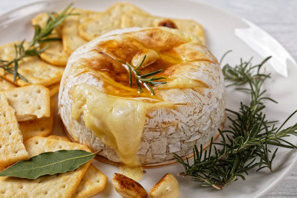 Baked camembert french soft cheese topped by olive oil with rosemary sprigs, garlic cloves, and bay leafs served with crackers on a white plate on a white wooden background, top view, close-up stock photo