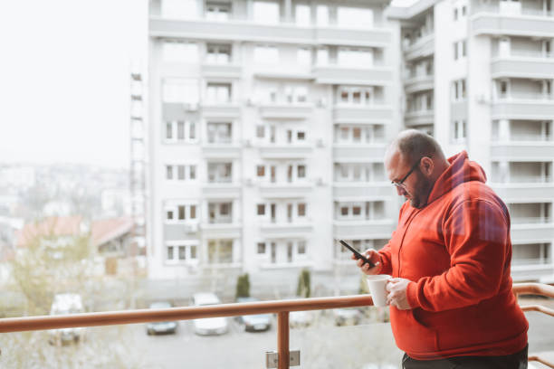 Bald Male Drinking Coffee On Balcony And Using Smartphone  fat man looks at the phone stock pictures, royalty-free photos & images