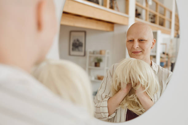 Bald Woman Smiling at Mirror Reflection portrait of bald adult woman looking in mirror holding wig while standing in warm-toned home interior, alopecia and cancer awareness, copy space wig stock pictures, royalty-free photos & images