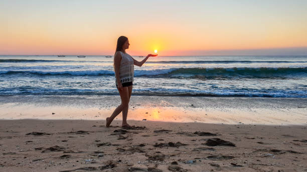 bangli - una ragazza che tiene un sole al tramonto in mano sulla spiaggia - forced perspective foto e immagini stock