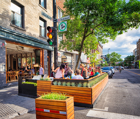 Montreal, Canada - July 29, 2016: People are enjoying Summer at a terrasse in front of a bar on the plateau's Mont-Royal avenue in Montreal on a late Summer afternoon. Others are walking on the sidewalk or biking in the streets. Yellow traffic light is on. A few cars are parked behind the terrasse.