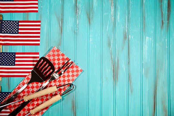 Happy Independence Day America!  Barbeque utensils on checked cloth. USA flags form a left side border with copyspace to right. Teal blue wooden beadboard background with grunge painted effects.  Memorial Day, July 4th, Veteran's Day, Labor Day, Flag Day concepts.  American pride and patriotism.