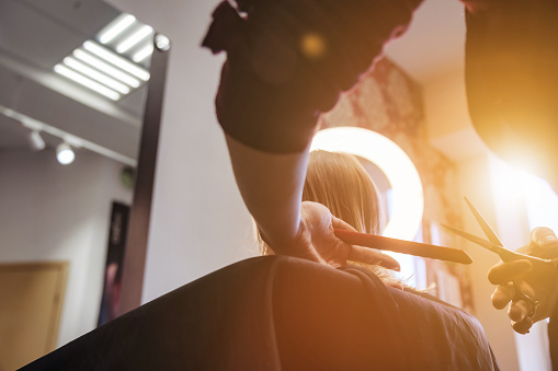 Back view hairdresser makes hairdo for baby in barber shop. Barber woman make fashionable pretty hairstyle for cute little blond girl child in modern barbershop. Hairstyles and beauty. Copy space