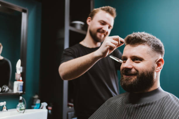 Barber young man doing haircut to happy bearded male customer stock photo