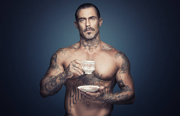 Bare chested man with tattoos holding a cup of tea. Bare chested adult caucasian man with religious tattoos holding scup of tea. Blue background. Vintage tea cup. gangster stock pictures, royalty-free photos & images