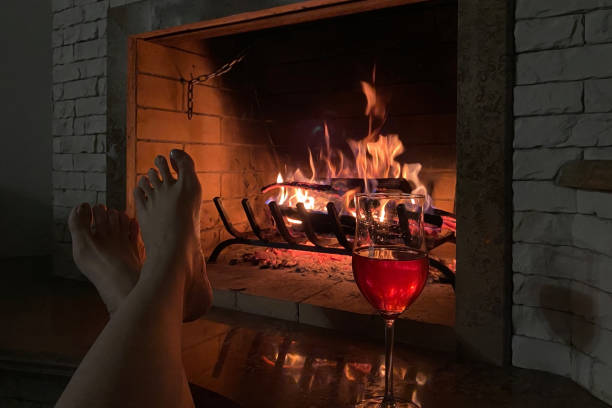 Barefoot woman resting in a cozy house with a glass of wine against the background of a burning fireplace, cozy warm moments stock photo
