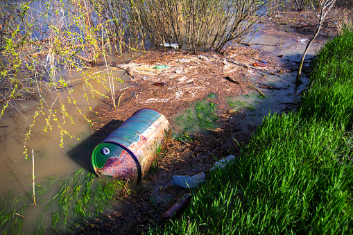 Barrel of toxic waste dumped into the river along with plastic bottles and other garbage polluting ecosystem.