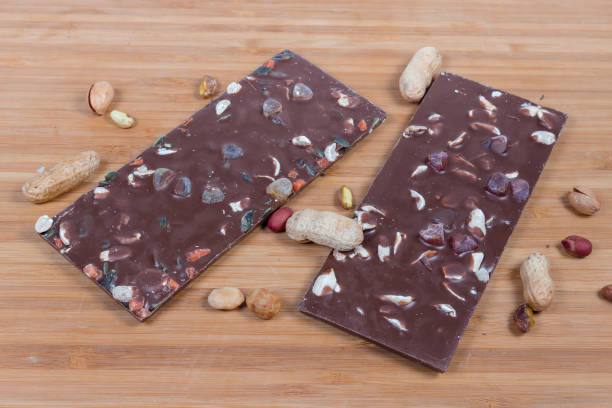 Bars of chocolate with nuts among the scattered various nuts Two bars of dark chocolate with different nuts and raisins among the scattered various nuts on the wooden surface semi sweet chocolate stock pictures, royalty-free photos & images