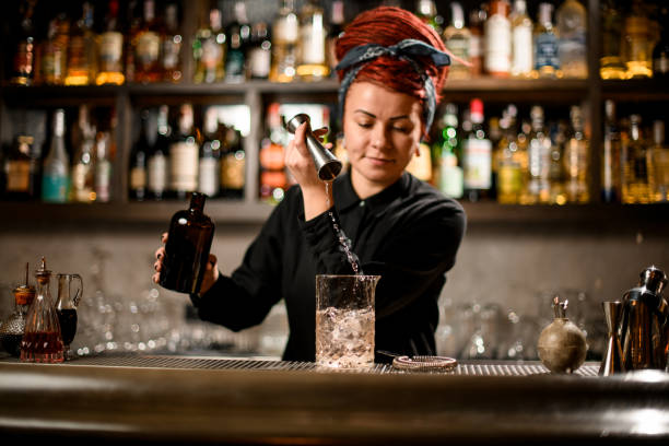 Bartender pours liquor from bottle with jigger Female bartender with ginger dreadlocks pours liquor from glass bottle with steel jigger bar drink establishment stock pictures, royalty-free photos & images