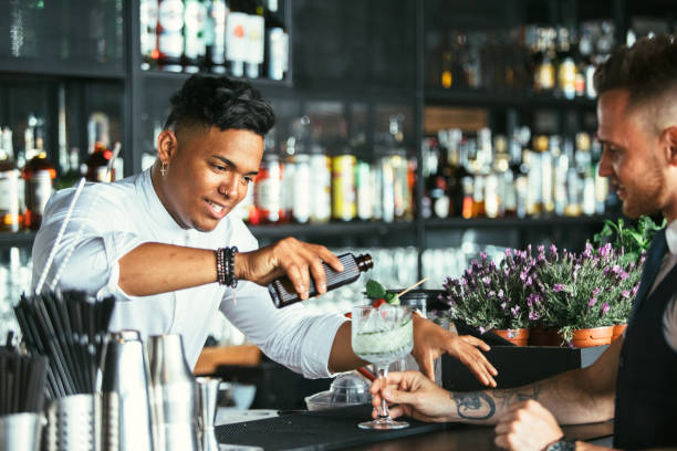Bartender prepares a cocktail to a waiter Mixed race male expert bartender is serving some tonic for a cocktail at the bar counter while a smiling waiter is holding the cocktail glass bartending stock pictures, royalty-free photos & images