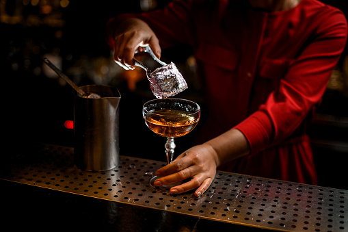 bartender woman's hand holding tongs with large ice cube over glass with drink on bar counter