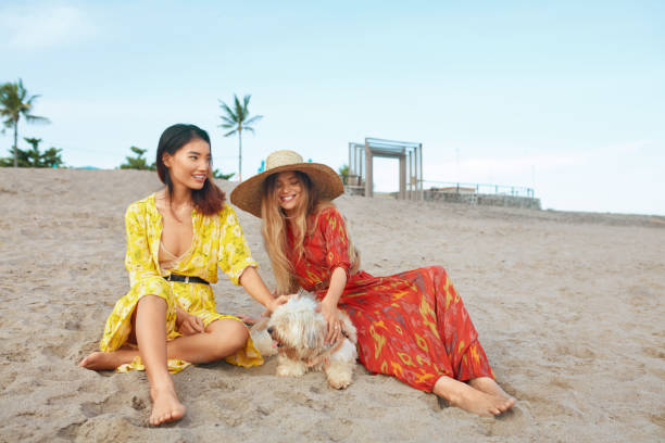 Beach. Women With Dog Sitting On Sandy Coast. Fashion Girls In Bohemian Clothing With Straw Hat Enjoying Resting On Tropical Shore. Boho Style For Fashionable Look And Walk On Resort. Beach. Women With Dog Sitting On Sandy Coast. Fashion Girls In Bohemian Clothing With Straw Hat Enjoying Resting On Tropical Shore. Boho Style For Fashionable Look And Walk On Resort. indonesian girl stock pictures, royalty-free photos & images