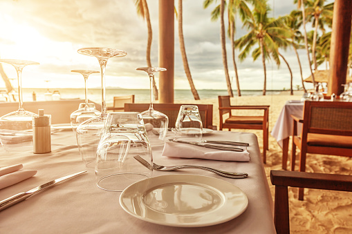 Photo of an outdoor dining table with place settings on a Fijian beach at sunset. Primary focus is on plate in the foreground.