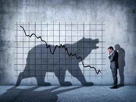 A businessman covers his eyes as he can't watch a descending stock chart and an ominous shadow of a bear that is cast on the wall above him.