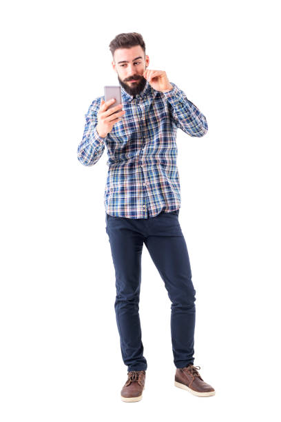 Bearded hipster man twirling mustache using mobile phone reflection as mirror Bearded hipster man twirling mustache using mobile phone reflection as mirror. Full body isolated on white background. mirror object photos stock pictures, royalty-free photos & images